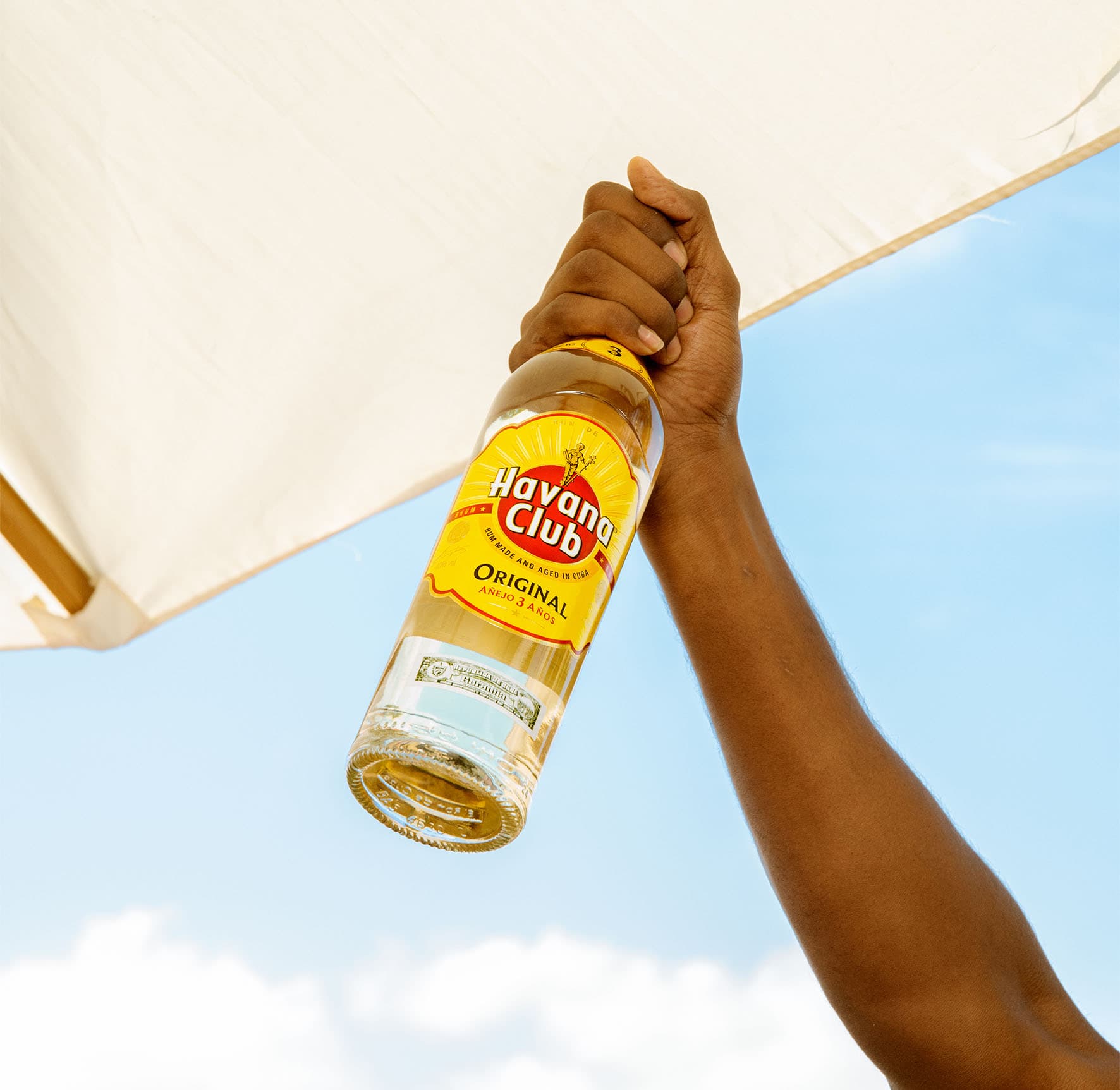 Hand hält eine Flasche Havana Club 3 Años Rum vor blauem Himmel und hellem Sonnenschirm.
