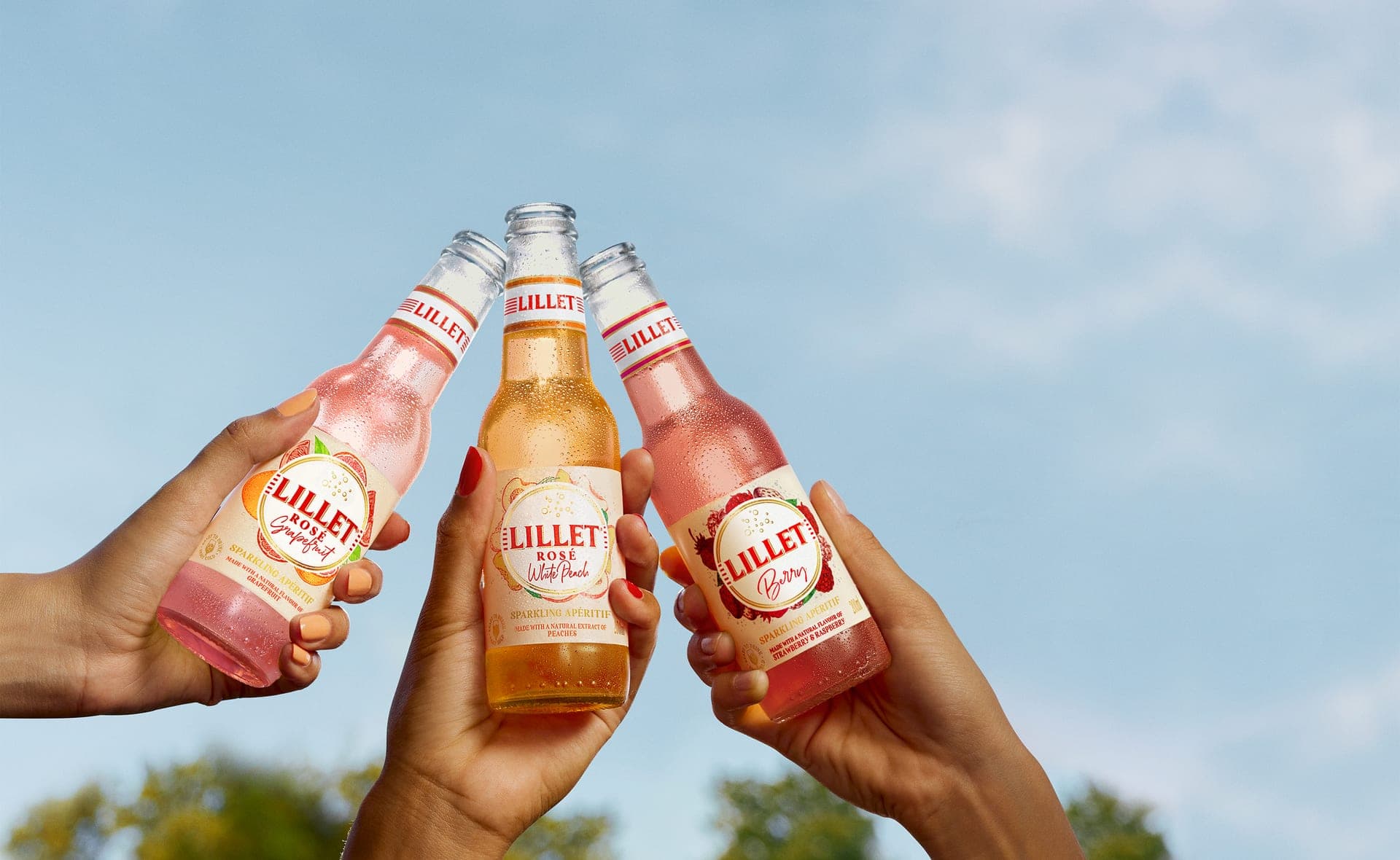 Drei Hände stoßen mit gekühlten Lillet Ready-to-Drink Flaschen in verschiedenen Sorten vor blauem Himmel an.