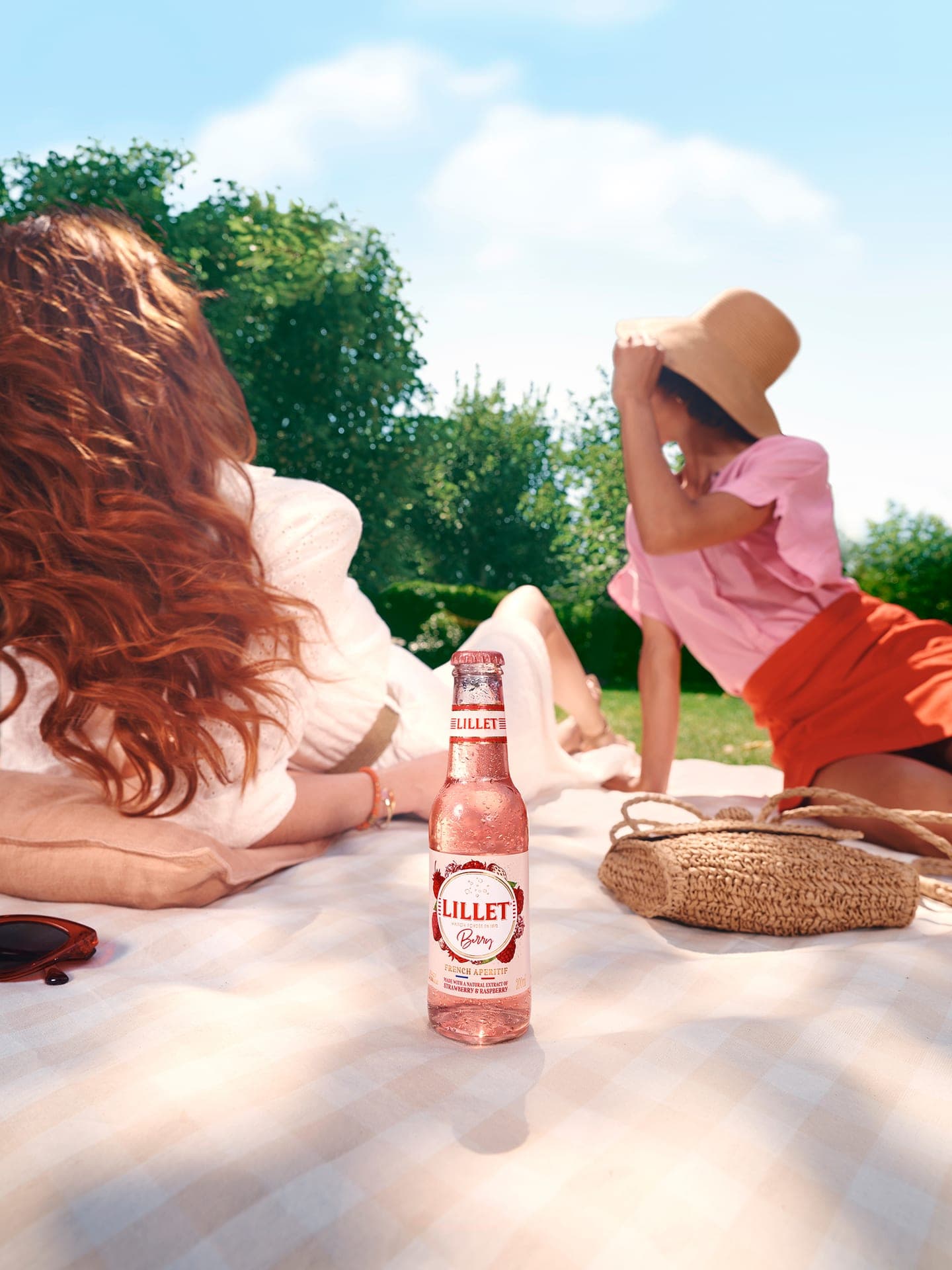 Gekühlte Flasche Lillet Berry auf einer Picknickdecke im sonnigen Garten, im Hintergrund entspannen zwei Personen im Freien.