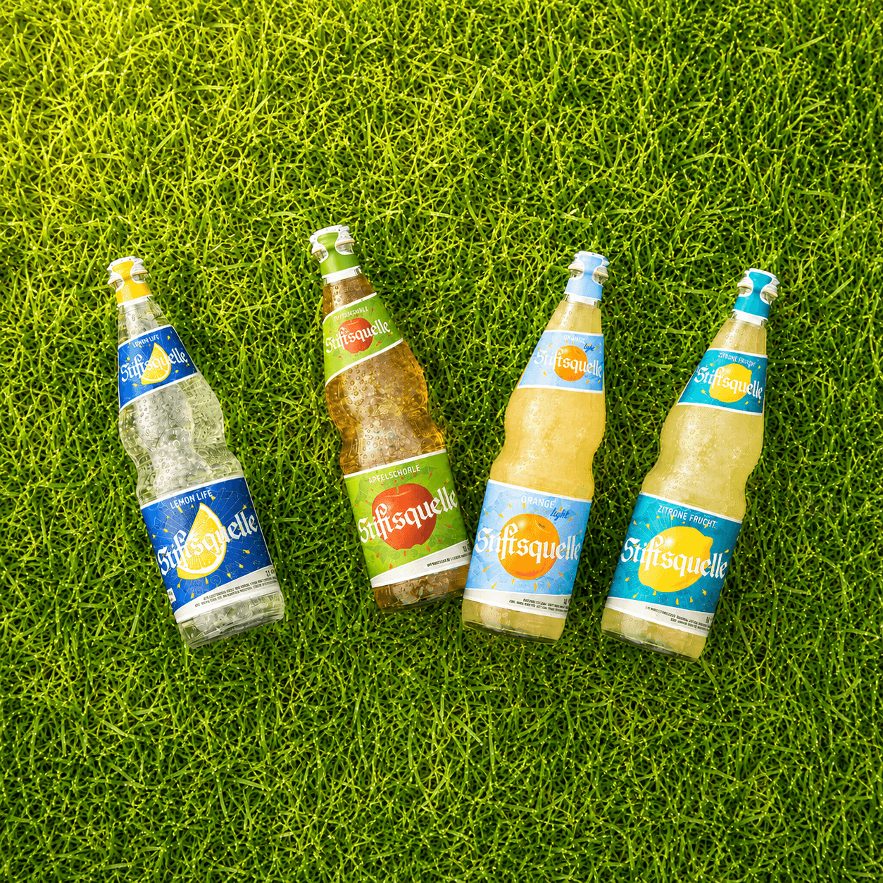 Vier Stiftsquelle-Glasflaschen mit Lemon Life, Apfelschorle, Orange light und Zitrone Frucht liegen gekühlt auf grünem Gras.